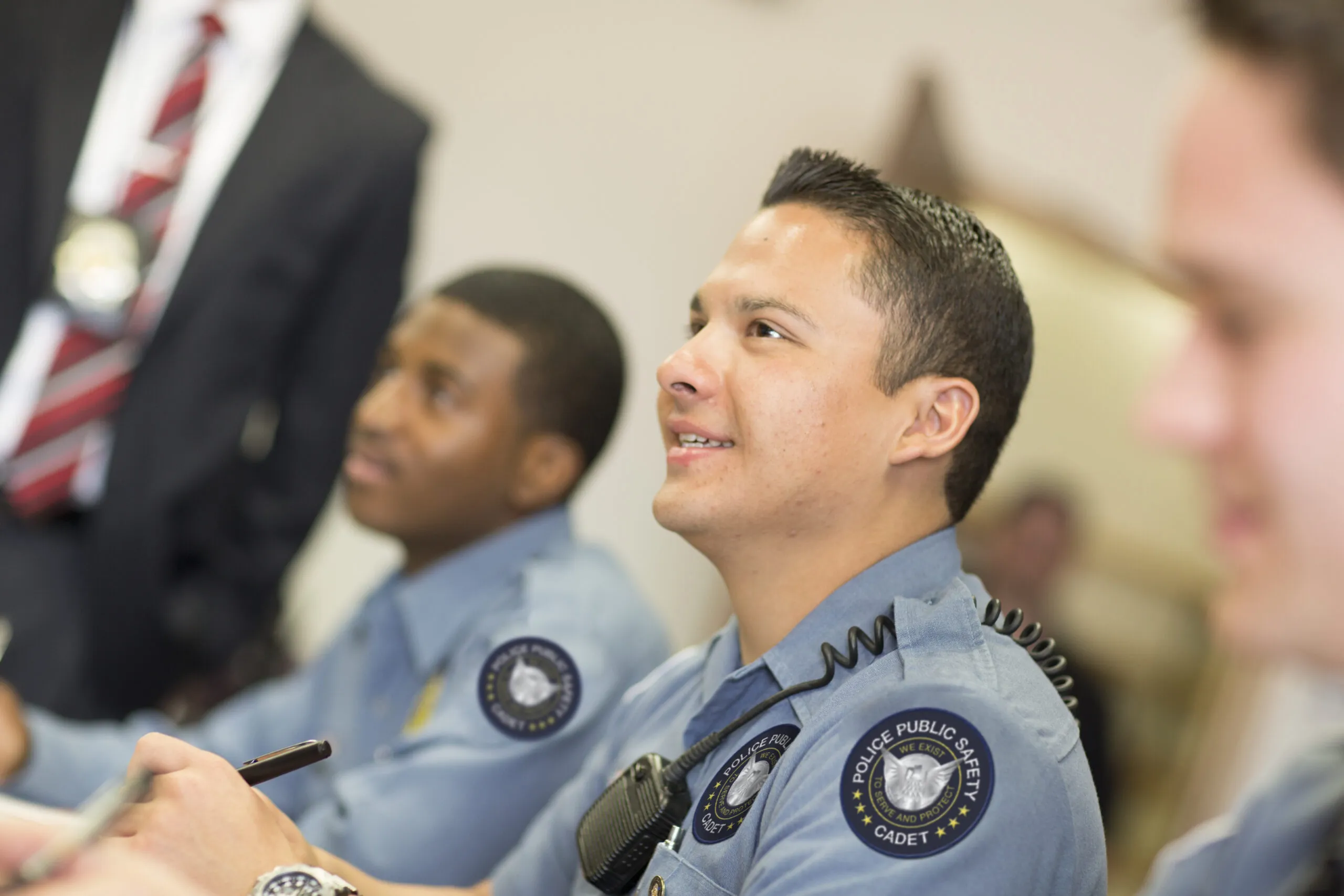 Two law enforcement officer sitting in a briefing type setting, both are wearing light blue shirts with patches and are smiling.