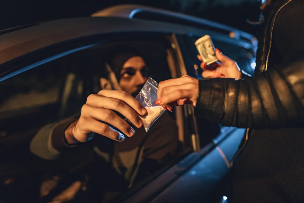 Male subject in vehicle exchanging money for a white substance in a baggie with only hands from the other person visible.